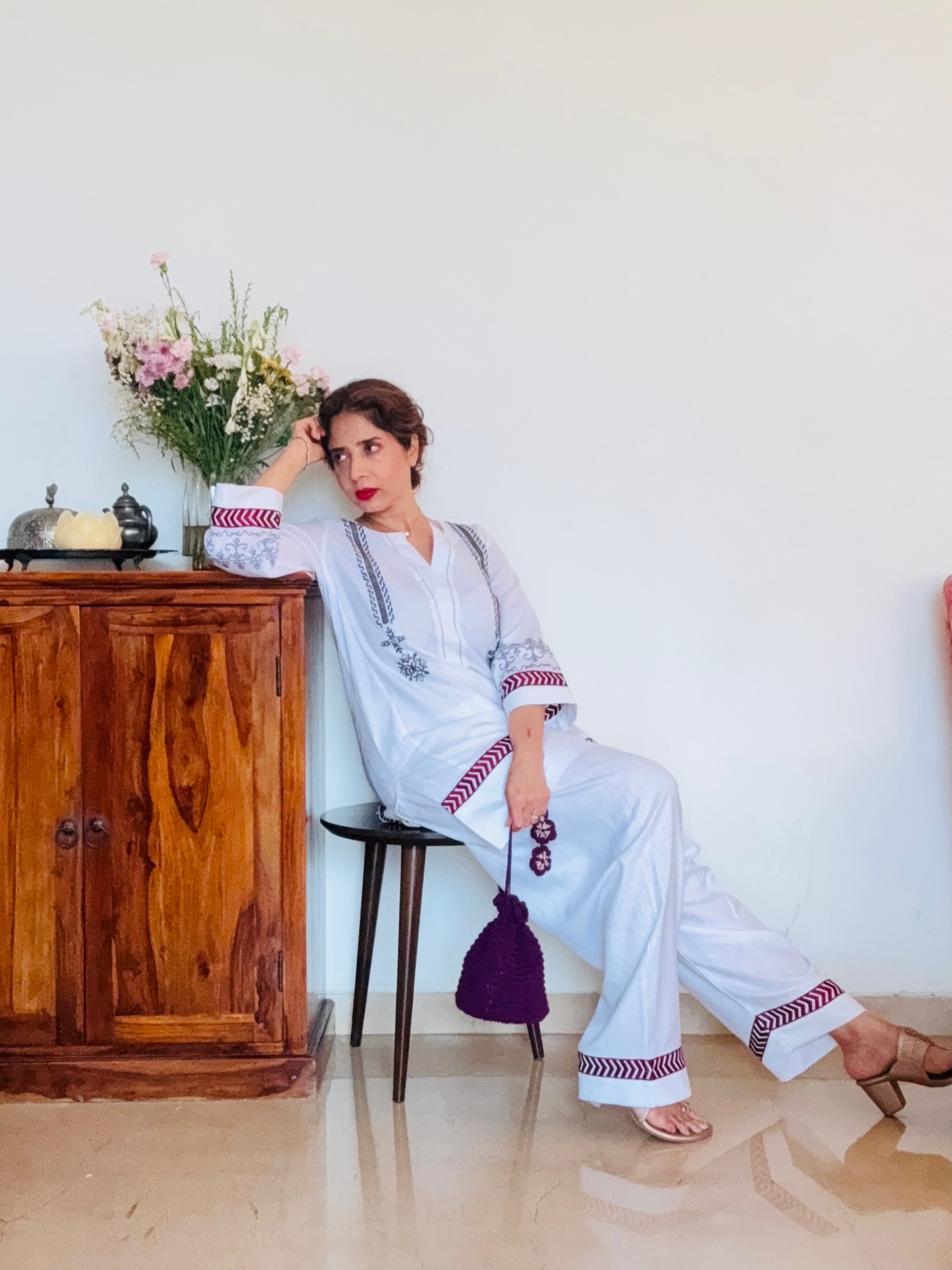 Aiana in a white hand-block co-ord seated beside a carved wooden cabinet with fresh flowers